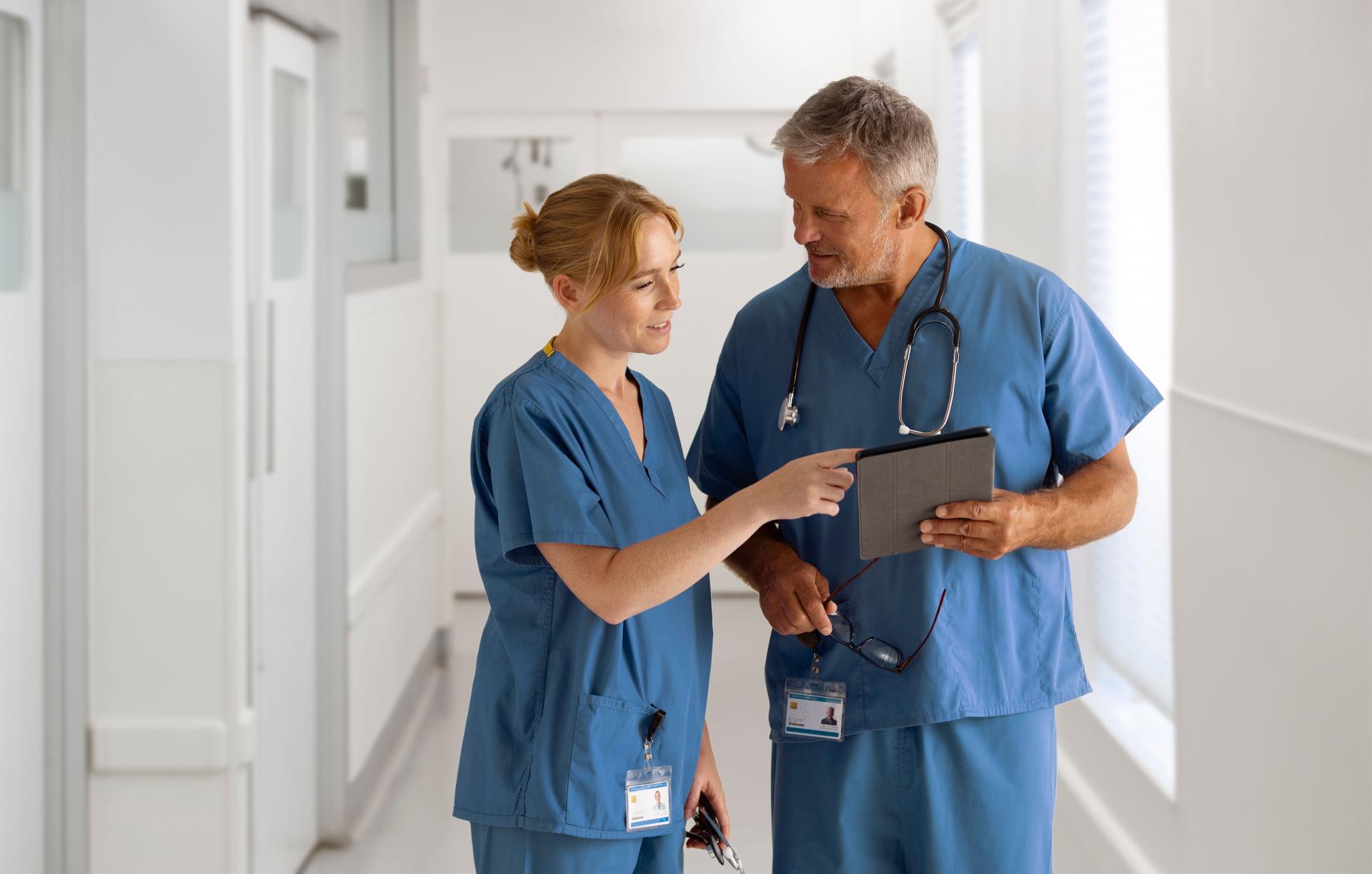 Medical Team Wearing Scrubs Meeting In Hospital Corridor Looking At Digital Tablet Together